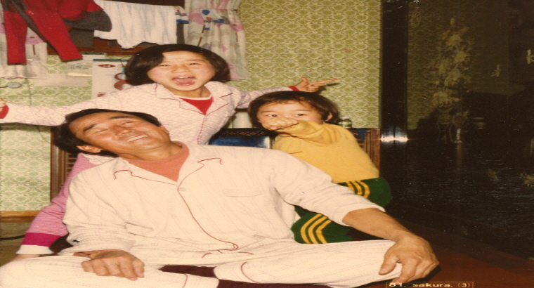 아버지는 앉아 웃고 있고, 유년시절 큰딸과 작은딸은 아버지의 뒤에서 개구진 모습으로 찍은 사진